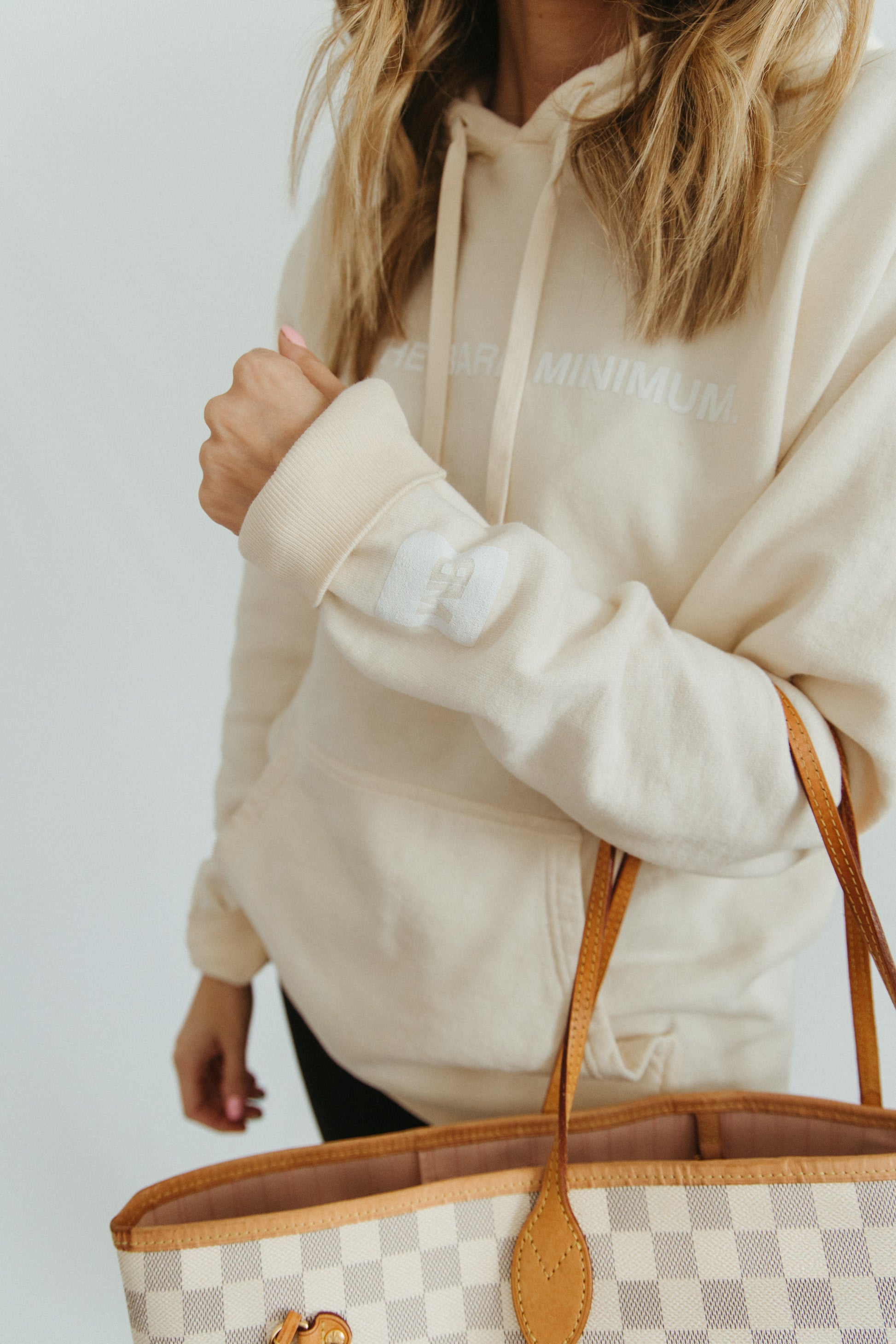 Bone white hooded sweatshirt with white writing that says 'Doing The Bare Minimum' across the chest; white logo on the sleeve above the wrist